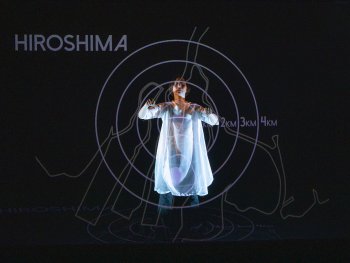 Tänzerin in weißer Kleidung, die in einer projizierten Grafik mit dem Wort 'HIROSHIMA', verschlungenen und geraden Linien und konzentrischen Kreisen steht, die die Karte einer Landschaft von oben betrachtet darstellen und den Explosionsradius einer Atombombe in Kilometern angeben.