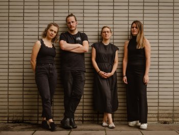 Drei Frauen und ein Mann stehen ganz in schwarz gekleidet vor einer Wand gelehnt und schauen in die Kamera.