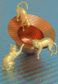 Drei goldfarbene Katzenfiguren um einen rot-gold gestreiften Hut auf blau-grün gestreiftem Untergrund.