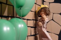 Puppe mit Krone vor einer Steinmauer mit Luftballons am Bildrand