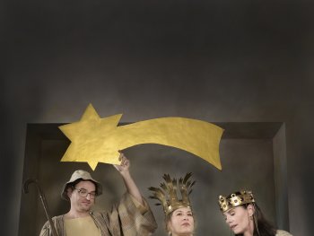 Vier Personen in gold- und cremefarbenen Gewändern, eine hält einen goldenen Stern mit Schweif, eine andere beugt sich über einen Korb mit Tuch und Holzspielzeug.