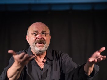 Copyright: Michl Zirk Michl Zirk, Erzähler, steht mit ausgebreiteten Händen auf einer Bühne, trägt ein schwarzes Hemd, der Hintergrund ist dunkel.