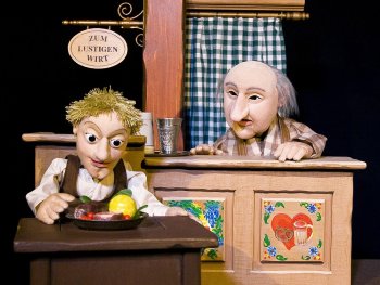 Foto, auf dem der Tischlerlehrling im Wirtshaus Essen auf seinem Tischlein deck dich herbeizaubert und vom gierigen Wirt dabei beobachtet wird.