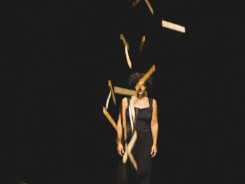 Foto: Maria Bayer
Frau mit lockigem Haar in schwarzem ärmellosem Kleid vor schwarzem Hintergrund, umgeben von fallenden Holzstäben.