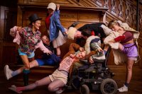 Sechs Personen in bunter Kleidung und verschiedenen Posen vor einer Holzvertäfelung, eine Person sitzt im Rollstuhl.