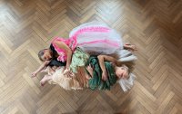 Drei Personen liegen eng aneinander auf einem Parkettboden, tragen bunte, fließende Kleider in Rosa, Grün und Beige.