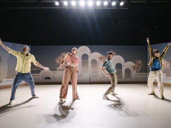 An der Bühnenkante zwei Männer und zwei Frauen in energetischen Posen, im Hintergrund Gegenlicht der Scheinwerfer, auf der Bühnenrückwand pastellfarbiges Fotomotiv (antike Bögen und Palmen)