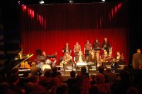 Jazzorchester auf Bühne mit roten Vorhängen, Musiker spielen verschiedene Instrumente wie Klavier, Schlagzeug und Blasinstrumente.