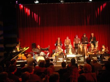 Foto: Sunday Night Orchestra
Jazzorchester auf Bühne mit roten Vorhängen, Musiker spielen verschiedene Instrumente wie Klavier, Schlagzeug und Blasinstrumente.