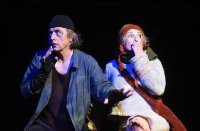 Zwei Schauspieler·innen halten sich erschrocken eine Hand vor den Mund. Links ein Mann in grauer Kleidung mit schwarzen Handschuhen und Mütze, dezent als Wolf geschminkt. Rechts eine Frau in flauschigem Wollpulli mit roter Mütze, das Schaf.