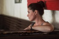 Frau mit hochgestecktem Haar und grauem Trägershirt lehnt mit Arm auf einem Klavier, rotes Holzpaneel im Hintergrund.