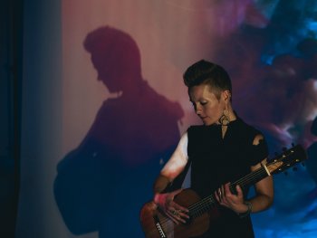 Foto: Andreas Haals
Person mit kurz geschnittenem Haar spielt akustische Gitarre vor einer Wand mit buntem Licht und Schatten.