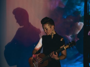 Person mit kurz geschnittenem Haar spielt akustische Gitarre vor einer Wand mit buntem Licht und Schatten.