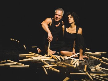 Foto: Maria Beyer
Zwei Personen in schwarzer Kleidung sitzen auf dem Boden, umgeben von verstreuten Holzstäben.