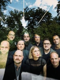 Gruppe von Menschen im Freien vor Bäumen, einige tragen dunkle Kleidung, eine Person mit blondem Haar und schwarzem Oberteil sitzt vorne.