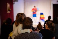 Ein Kind ist auf dem Arm des Vaters, sie sind von hinten zu sehen. Sie schauen auf die Bühne, wo man verschwommen eine bunt gekleidete Person erkennen kann.