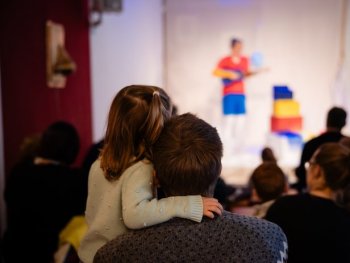 Ein Kind ist auf dem Arm des Vaters, sie sind von hinten zu sehen. Sie schauen auf die Bühne, wo man verschwommen eine bunt gekleidete Person erkennen kann.