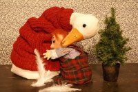 Eine weiße Gans mit rotem Pullover beugt sich über eine kleine Puppe mit rotem Haar, die ein kariertes Kleid trägt. Neben der Puppe steht ein kleiner, grüner Tannenbaum in einem schwarzen Topf. Im Hintergrund ist eine rot braun gemusterte Wand.