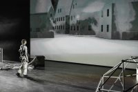 Person in heller Kleidung steht in einem dunklen Raum vor einer großen Wandprojektion mit Häusern, alles ist in ein graues Licht getaucht