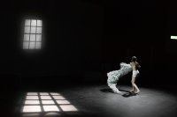 Person in Unterwäsche bewegt sich auf einer dunklen Bühne mit projizierten Fenstermustern auf Boden und Wand. Sie schwingt ein helles Tarnnetz um sich.