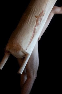 Teilweise sichtbarer nackter Mann trägt ein großes, längliches, helles Holzobjekt mit zwei Griffen vor dem Körper, schwarzer Hintergrund.