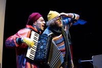 Zwei Schauspieler in farbenfrohen Kostümen, einer spielt Akkordeon