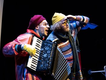 Zwei Schauspieler in farbenfrohen Kostümen, einer spielt Akkordeon