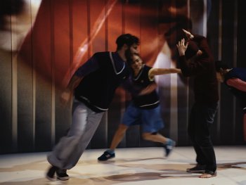 Fünf Personen in sportlicher Kleidung bewegen sich dynamisch auf einer Bühne mit rot-braunem Hintergrund und weißen Bodenmarkierungen.