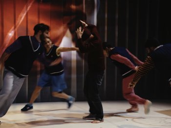 Fünf Personen in sportlicher Kleidung bewegen sich dynamisch auf einer Bühne mit rot-braunem Hintergrund und weißen Bodenmarkierungen.