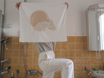 Person hält ein mit einem Schädel in Weiß und Beige besticktes Tuch in die Höhe, sodass das Gesicht der Person verdeckt wird