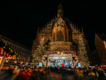 Copyright: Kristof Göttling Weihnachtlich beleuchtete Bühne mit mehreren Personen vor der beleuchteten Fassade der Nürnberger Frauenkirche, umgeben von großen Weihnachtsbäumen.