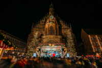 Weihnachtlich beleuchtete Bühne mit mehreren Personen vor der beleuchteten Fassade der Nürnberger Frauenkirche, umgeben von großen Weihnachtsbäumen.