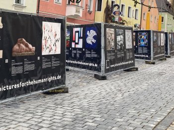 Copyright: Pictures fpr the Human Rights e.V. An Bauzäunen aufgehängte Banner zeigen Bilder und Text zu der Allgemeinen Erklärung der Menschenrechte in einer Fußgängerzone.