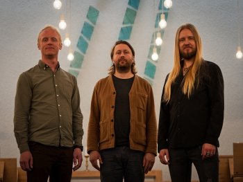 Drei Personen stehen in einem Raum mit hohen Wänden, hängenden Glühbirnen und Holzbänken im Hintergrund.