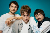 Drei junge Drei junge Männer in lässiger Kleidung vor blauem Hintergrund, einer trägt ein graues Hemd über weißem T-Shirt.