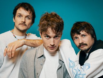 Drei junge Drei junge Männer in lässiger Kleidung vor blauem Hintergrund, einer trägt ein graues Hemd über weißem T-Shirt.