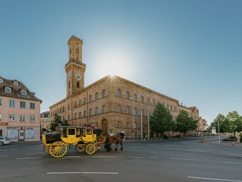 Gelbe Postkutsche mit zwei Pferden vor dem historischen Rathaus mit hohem Turm in der Sonne.