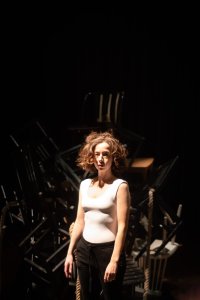 Frau mit lockigem Haar in weißem Oberteil und dunkler Hose steht vor einem Haufen übereinander gestapelter Stühle in dunklem Raum.