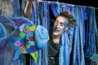 Eine als Meereswesen geschminkte Person blickt zwischen blauen Stoffbändern auf eine bunte Fisch-Puppe herab.