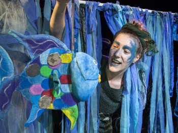 Eine als Meereswesen geschminkte Person blickt zwischen blauen Stoffbändern auf eine bunte Fisch-Puppe herab.