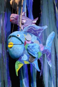 Bunt bemalte Fisch-Stabpuppen mit glänzenden Schuppen schweben vor blauen Stoffbahnen als Bühnenkulisse.