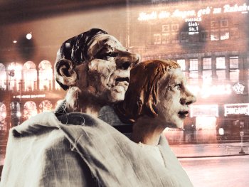 Zwei Figuren - im Hintergrund, die beleuchtete Hauswand einer Stadt
