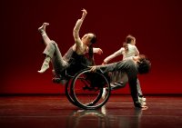 Drei Tänzer auf Bühne vor rotem Hintergrund, einer davon in Rollstuhl, alle in dynamischer Pose mit ausgestreckten Armen.