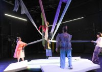 Person in rotem Oberteil sitzt auf einem weißen Tuch, das von mehreren Personen gehalten wird, auf einer weißen Bühne in dunklem Raum