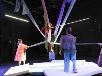 Foto: Jörg Landsberg
Person in rotem Oberteil sitzt auf einem weißen Tuch, das von mehreren Personen gehalten wird, auf einer weißen Bühne in dunklem Raum