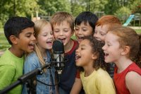7 Kinder singen oder sprechen draußen vor Spielplatz ins Mikrofon