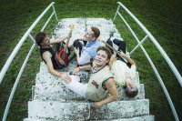 Vier Personen sitzen auf einer steinernen Treppe im Freien, zwei halten E-Gitarren, alle tragen Armbänder und lässige Kleidung.