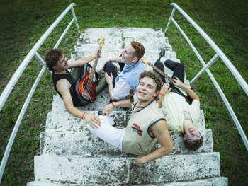 Vier Personen sitzen auf einer steinernen Treppe im Freien, zwei halten E-Gitarren, alle tragen Armbänder und lässige Kleidung.