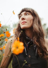 Frau mit langem, gewelltem Haar trägt ein ärmelloses, schwarz-weiß gepunktetes Hemd mit Knöpfen und einem gemusterten Halstuch, umgeben von orangefarbenen Blumen in einer Wiese