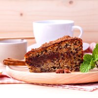 ein Stück Kuchen auf einem Holzteller, dahinter eine weiße Tasse auf Unterteller